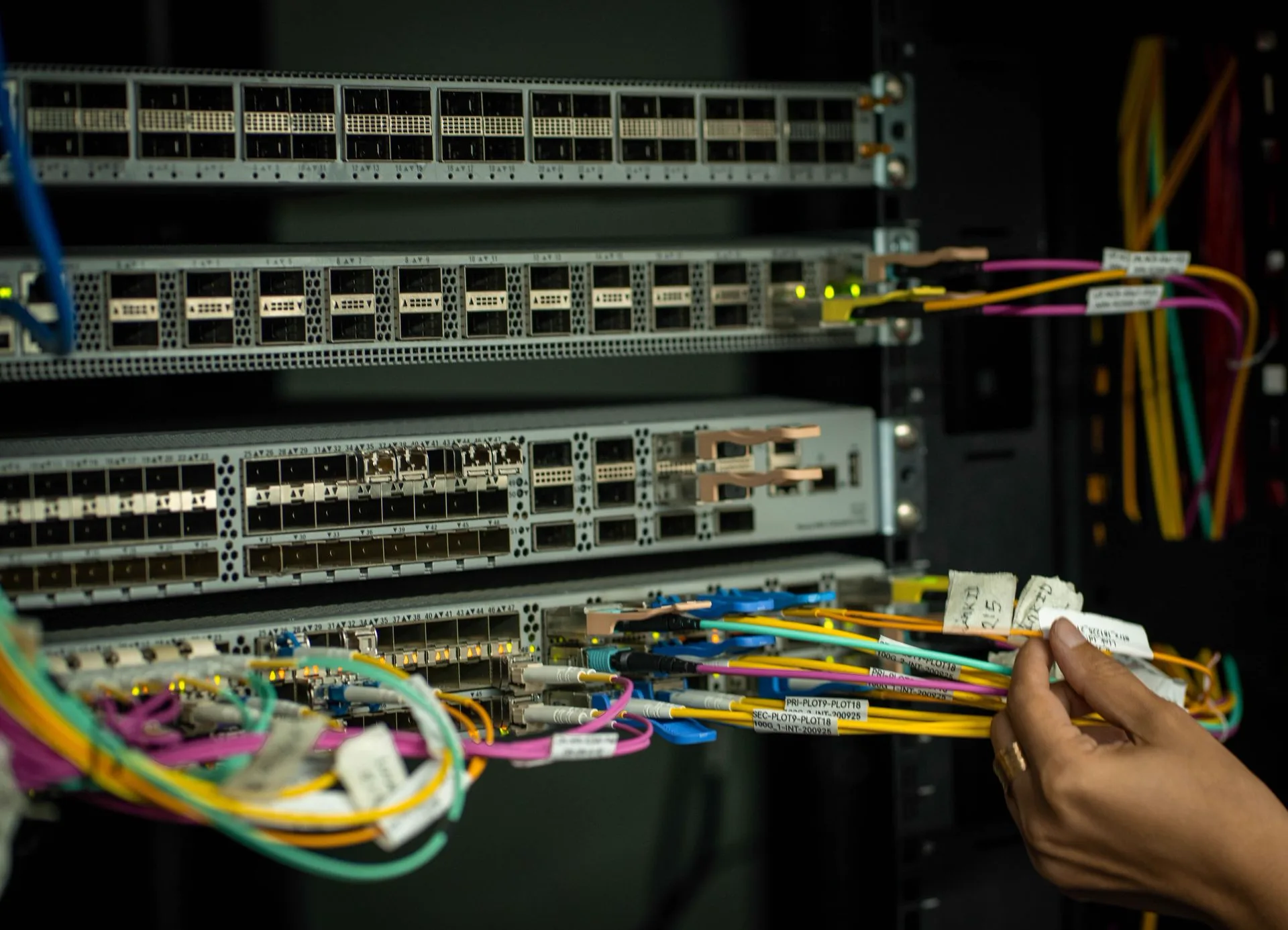 An engineer's hands routing labelled multicoloured fibre leads into a rack-mounted network switch, working under even ambient light in a live equipment room.