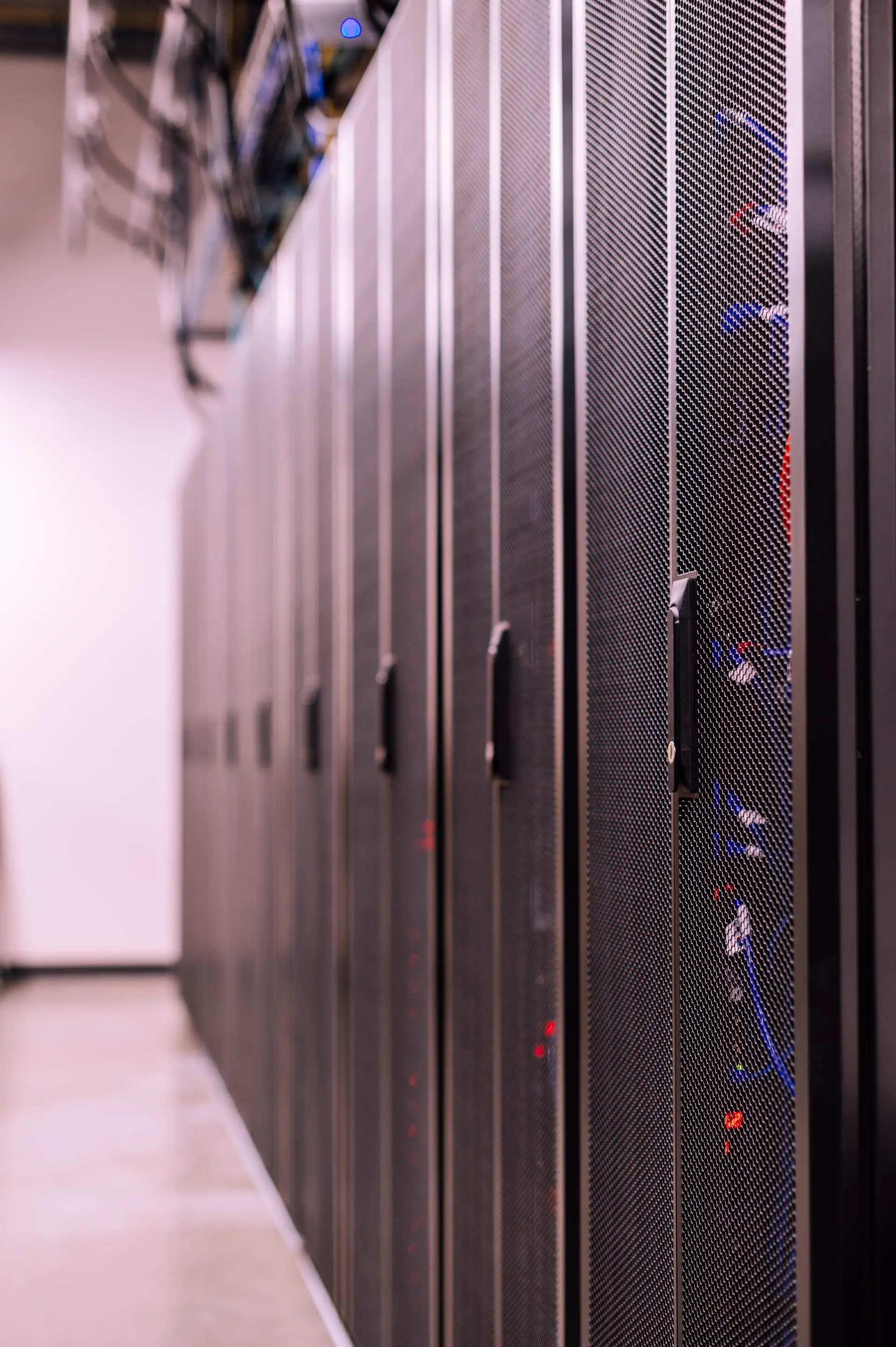 A long row of black perforated server cabinets running the length of a data centre aisle, status LEDs glowing red and blue behind the mesh doors.